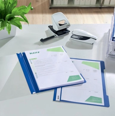Das Bild zeigt zwei blaue Leitz Plastik Hefter auf einem Schreibtisch, in denen Dokumente abgeheftet sind. Ein Symbol weist darauf hin, dass die Hefter aus 30 % recyceltem Kunststoff bestehen. Daneben stehen ein Locher sowie ein Tacker. Im Hintergrund ist eine Pflanze zu sehen.