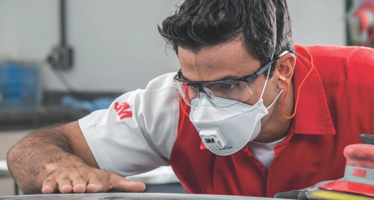 Mann mit Schutzbrille und Atemmaske von 3M inspiziert Oberfläche in einer Werkstatt.