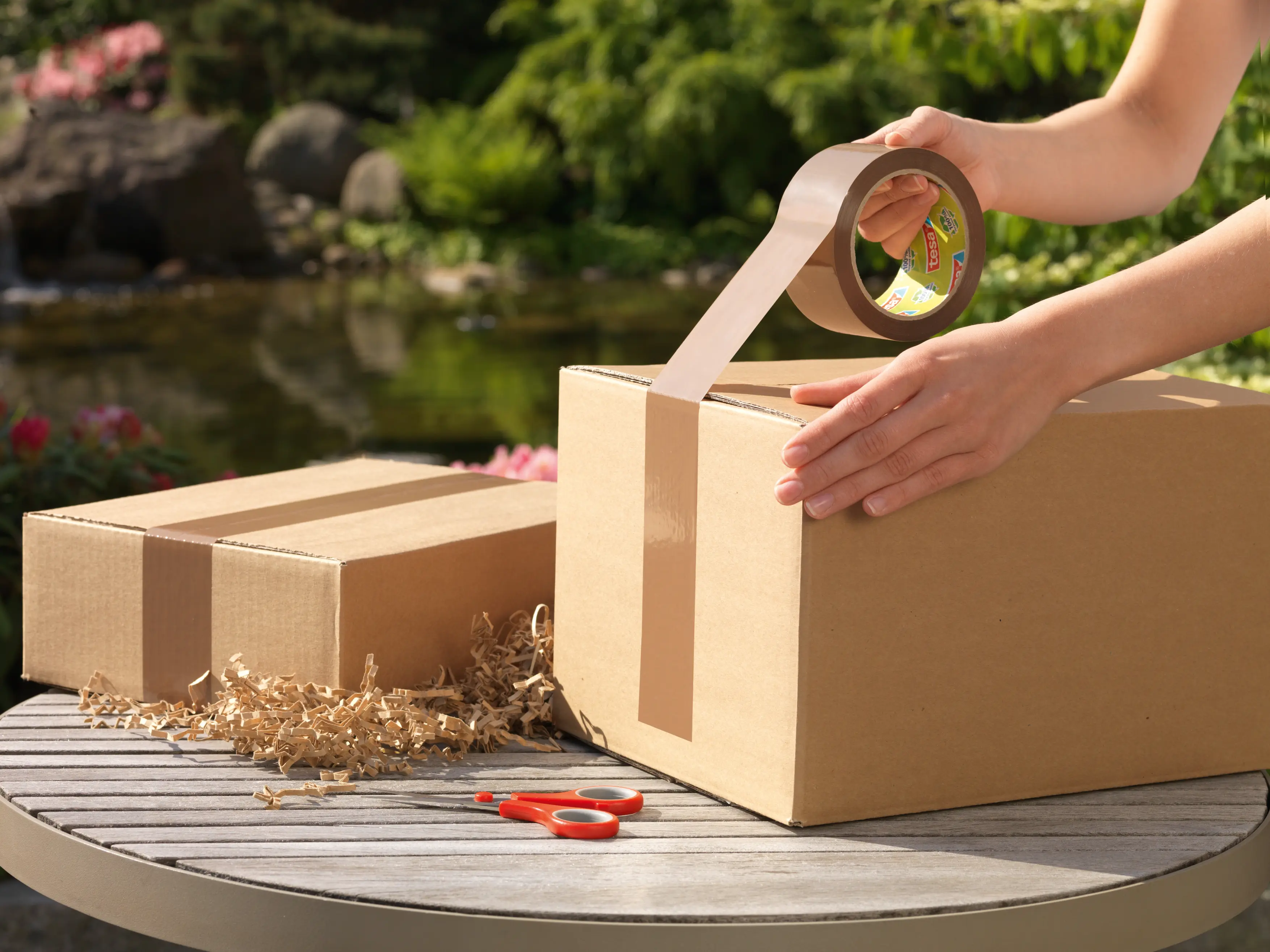 Die Abbildung zeigt eine Person, die im Freien auf einem Tisch ein Paket mit einem tesa Packband verschließt. Auf dem Tisch liegt eine Schere und Polstermaterial aus Papier.