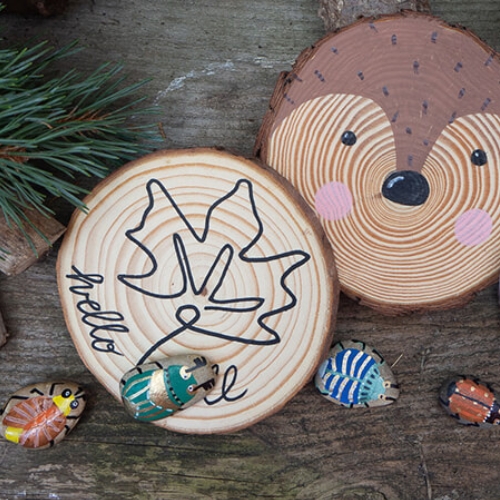 Drei bemalte Baumscheiben auf rustikalem Holz: Ahornblatt, Fuchs und Emaille-Tasse mit Camping-Motiv. Davor bemalte Steine als Käfer, umgeben von Holzscheiten und Kiefernzweigen."