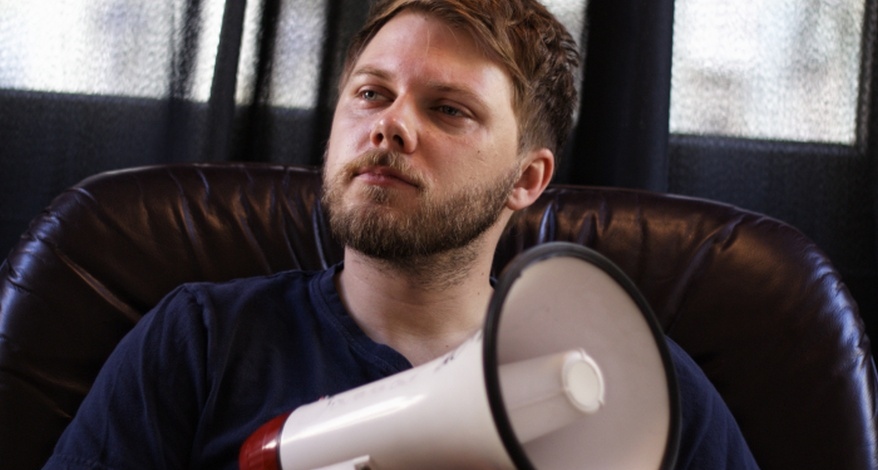 Ein Portrait des Poetry Slammers Christian Ritter. Er sitzt in einem Sessel aus Leder und hält ein Megaphone in der Hand. Im Hintergrund sind unscharf Fenster und dunkle Vorhänge zu erkennen.