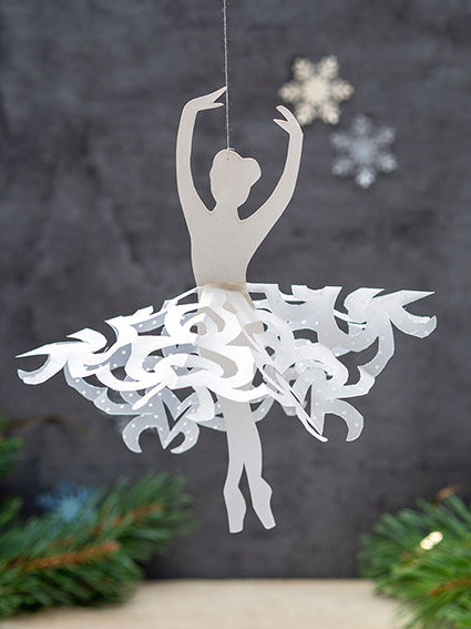 Vor einem dunklen Hintergrund hängt eine Ballerina aus weißem Papier von Artoz. Das Tutu ist aus Transparentpapier mit einer Scherenschnitttechnik zur Schneeflocke gearbeitet. Im Hintergrund hängen weitere Papierschneeflocken. Einige Tannenzweige dienen als zusätzliche Deko. Foto: Johanna Rundel