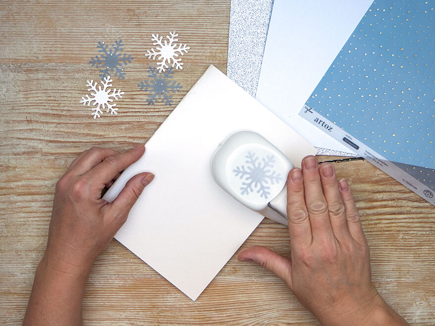 Jemand stanzt mit einem Motivstanzer Schneeflocken aus verschiedenen Papieren von Artoz als Streudeko aus. Der Untergrund ist ein rustikales, helles Holzbrett. Foto: Johanna Rundel