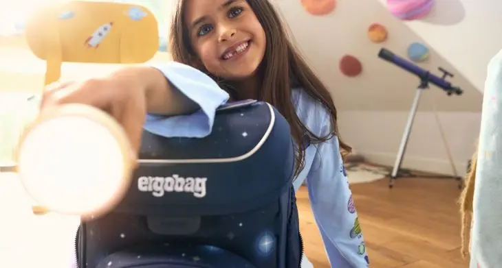 Ein Mädchen sitzt neben einem Schulranzen von ergobag und leuchtet mit einer Taschenlampe in die Kamera