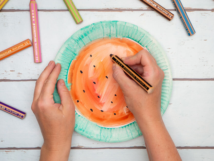 Jemand malt mit einem schwarzen Buntstift Punkte auf einen Pappteller, der wie eine Wassermelone angemalt ist: innen rot, der Rand grün. Der Untergrund ist hell. Foto: Johanna Rundel