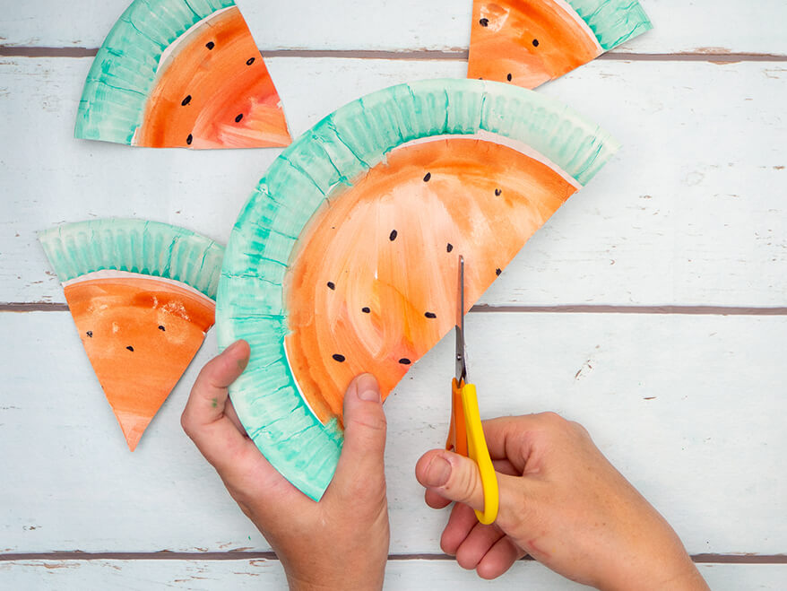 Jemand schneidet mit einer Schere von Staedtler einen Pappteller, der wie eine Wassermelone angemalt ist, in gleichgroße Dreiecke. Der Untergrund ist hell. Foto: Johanna Rundel