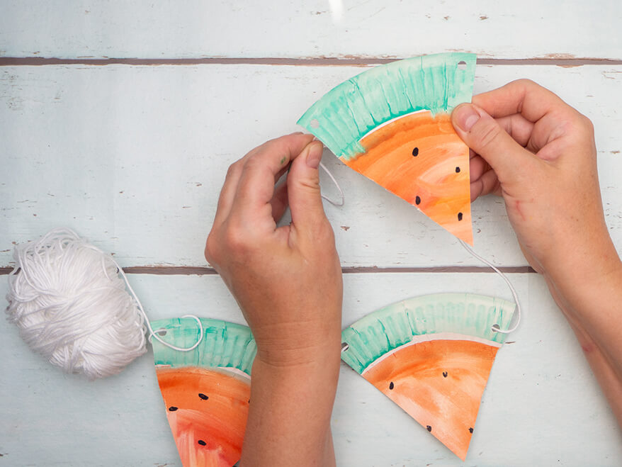 Jemand fädelt Dreiecke aus Papptellern auf ein weißes Stück Garn. Die Dreiecke sehen aus wie Wassermelonenstücke. Es wird eine Wassermelonen-Girlande. Der Untergrund ist hell. Foto: Johanna Rundel