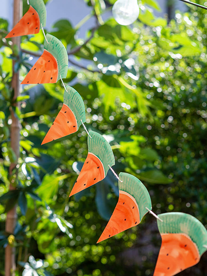 Eine Girlande aus Wassermelonen-Dreiecken aus Papptellern hängt in einem üppig grünen Garten. Foto: Johanna Rundel