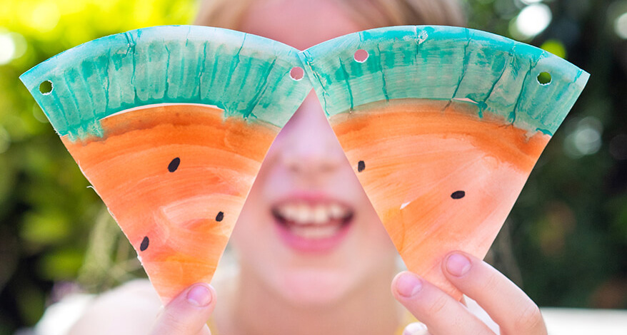 Ein Mädchen im Grundschulalter hält zwei Wassermelonen-Dreiecke aus Papptellern von ihr Gesicht und lacht dabei. Im Hintergrund ist das verschwommene Grün eines Gartens sichtbar. Foto: Johanna Rundel