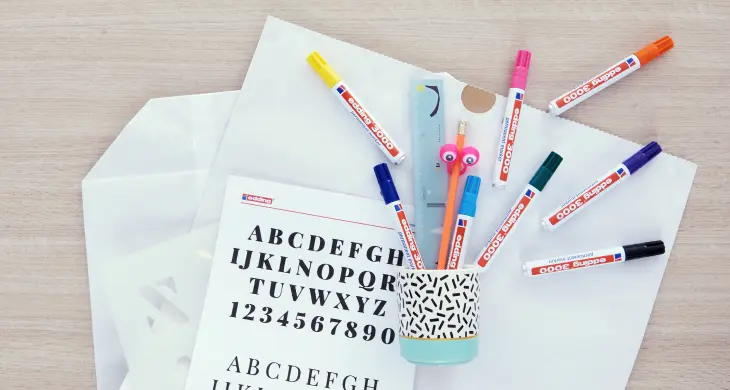 Bild mit einer Tasse und vielen edding Permanentmarkern 3000, die auf einem Blatt Papier liegen, auf dem das Alphabet in verschiedenen Strichstärken abgebildet ist.