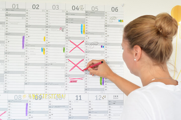 Man sieht den Rücken einer jungen Frau, die gerade mit einem roten Stift ein Kreuz in einen großen Wandkalender zeichnet. 