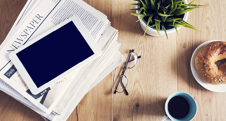 Blick aus der Vogelperspektive auf einen Tisch mit Tablet, Tasse, Brille, Zeitung und Pflanze.