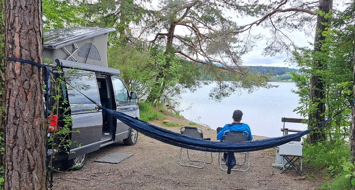 Person entspannt am See mit Camper, Hängematte und Campingstuhl.