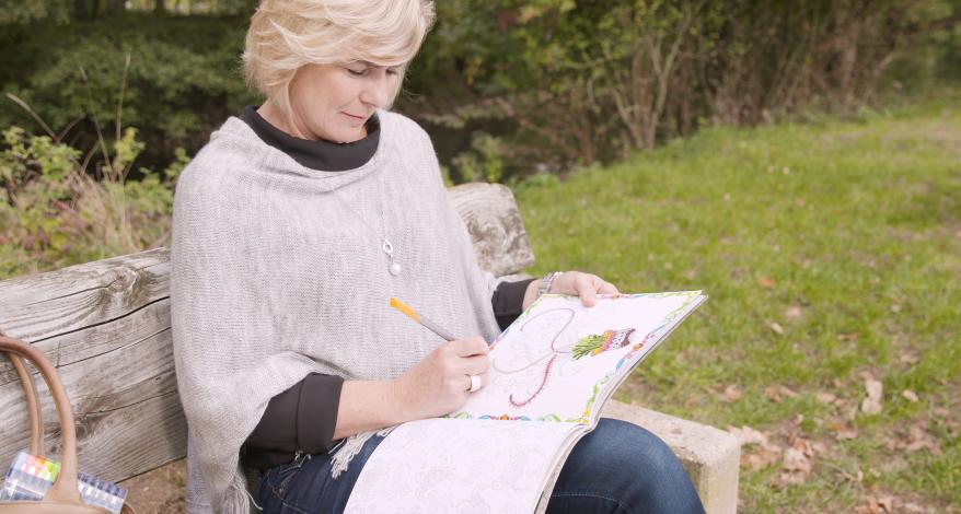 Eine Frau sitzt in der Natur und malt in einem Malbuch für Erwachsene.