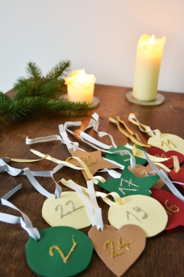 Viele bunte selbstgebastelte Adventskalender Anhänger in unterschiedlichen Formen liegen auf einem Tisch. Im Hintergrund stehen zwei Kerzen und es liegt ein Ast auf dem Tisch.