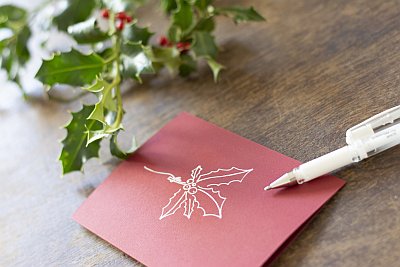 Hand zeichnet mit weißem Stift einen Mistelzweig auf rote Karte, daneben Ilex-Zweig.