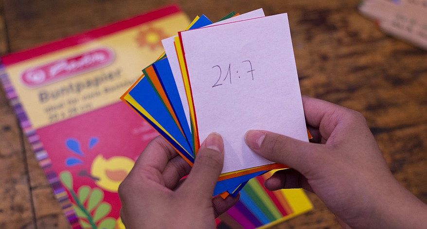 zwei Hände halten verschiedene Karten aus Buntpapier. Diese wurden mit Rechenaufgaben beschriftet. Der oberste Zettel ist weiß und wurde mit "21:7" beschriftet. Im Hintergrund ist unscharf ein Holztisch zu sehen, auf den ein Block mit Buntpapier von Herlitz liegt.