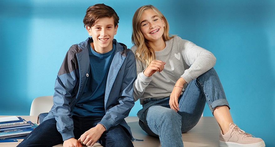 Zwei Jugendliche lachen und sitzen entspannt auf einem Tisch vor blauem Hintergrund.