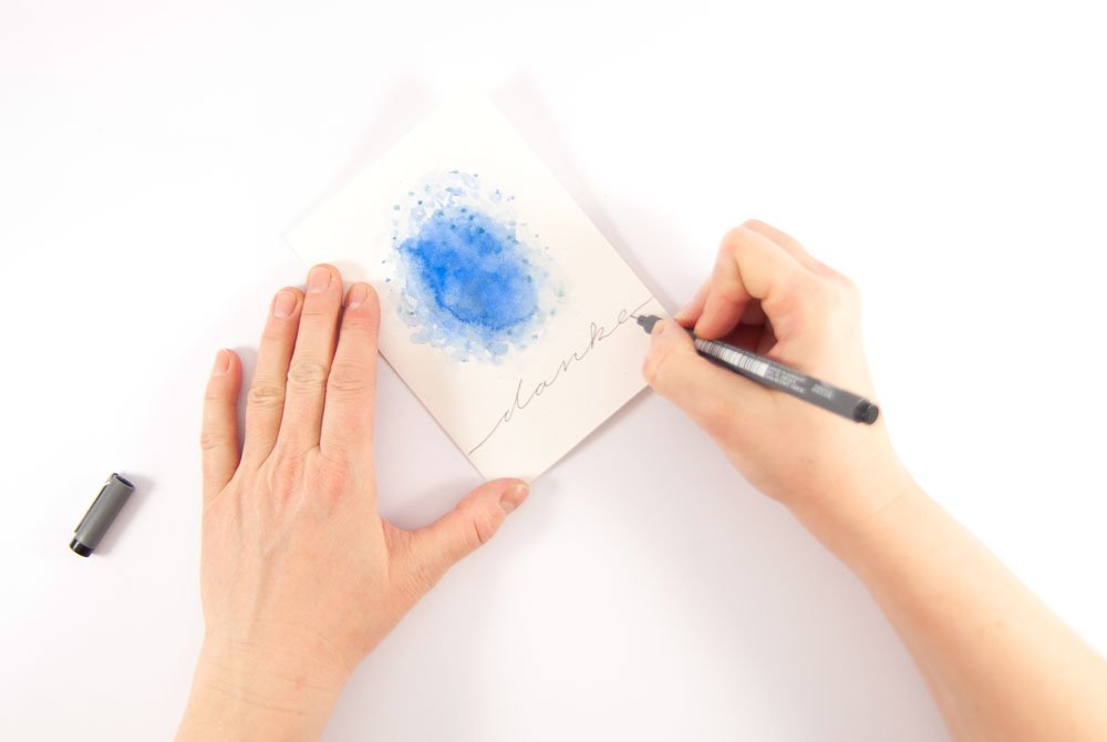 Hand schreibt „danke“ in Schreibschrift auf eine selbst gestaltete Dankeskarte.
