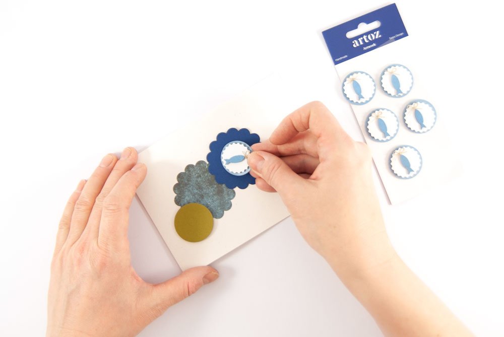 Hand klebt ein runden Aufkleber mit Fischmotiv auf eine selbst gestaltete Dankeskarte.