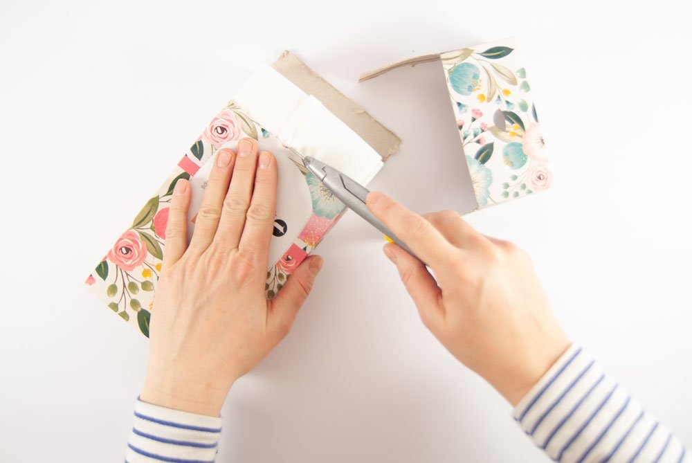 Auf einem weißen Untergrund liegt eine Pappschachtel, die von einer Hand gehalten wird. Mit der anderen Hand wird mit Hilfe eines Cutters ein Teil des Pappkartons abgeschnitten. Der Deckel wurde bereits abgeschnitten und liegt im oberen Bildrand.