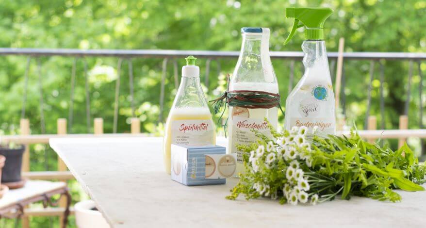 Selbstgemachte DIY-Haushaltsmitel (DIY-Spülmittel, DIY-Waschmittel und DIY-Badreiniger) stehen aufgereiht auf einem Tisch. Im Vordergrund liegt ein Blumenstrauß und Sticker von der Rolle von Avery Zweckform.