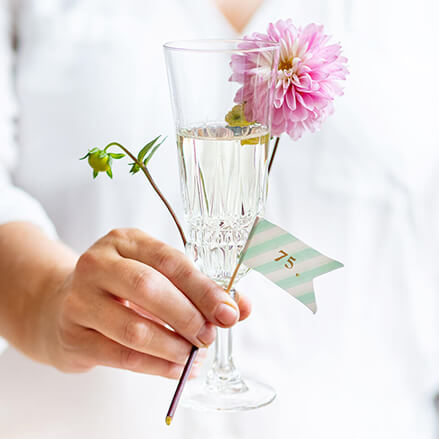 Eine Person hält ein Sektglas, eine Blume und einen Stab mit einer Wimpel.