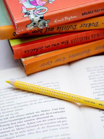 Auf einem aufgeschlagenen Buch liegt ein gelber Jumbo Grip Stift von Faber-Castell. Dahinter ist ein Stapel Bücher mit orangefarbenen Rücken. Foto: Johanna Rundel