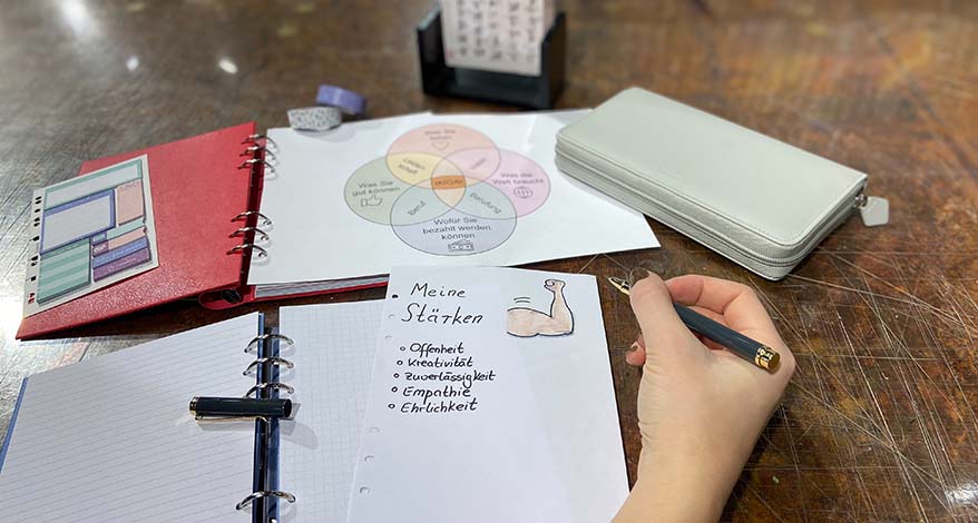 Eine Hand hält einen Kugelschreiber der Marke Filofax und schreibt auf ein Blatt Papier auf dem "Meine Stärken" steht. Im Hintergrund sieht man ein Notizbuch und einen Planner der Marke Filofax sowie ein Bild und Washitape.