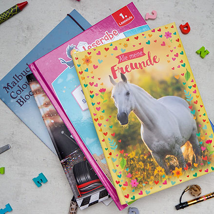 Auf einem hellgrauen Untergrund liegt ein Stapel mit Erstlesebüchern, Ausmalheften und Freundebüchern. Drumherum sind Stifte und kleine, bunte Radiergummis in Buchstabenform verteilt. Foto: Johanna Rundel