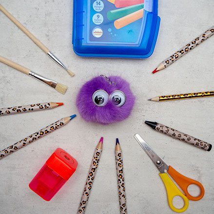 Auf einem hellgrauen Untergrund liegt ein Schlüsselanhänger in Form eines kleinen lila Fellpuschels mit Augen von ergobag. Diese Anhänger nennen sich Hangies. Drumherum liegen im Kreis verschiedene Stifte, eine Schere, ein Spitzer, Pinsel sowie eine Packung Schulknete. Foto: Johanna Rundel