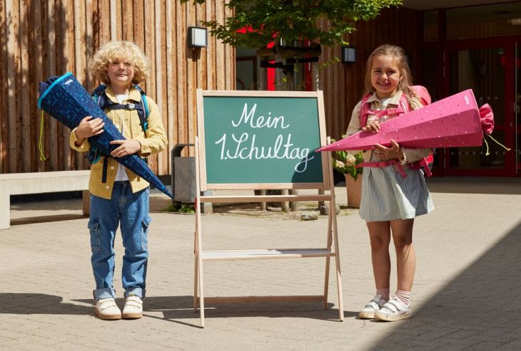 Zwei Kinder am ersten Schultag vor der Schule mit Schultüten in der Hand. Zwischen ihnen steht eine Tafel mit der Aufschrift "Mein 1. Schultag".