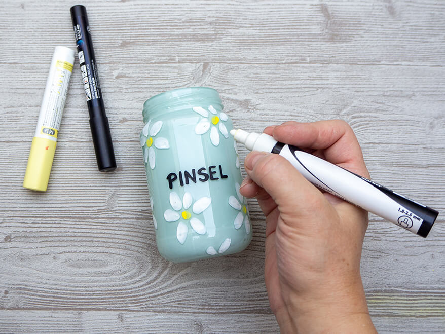 Jemand malt auf einem hellgrauen Holzuntergrund ein leeres Marmeladenglas, das von innen mit hellblauer Acrylfarbe ausgesprüht wurde, mit Acrylmarkern an.