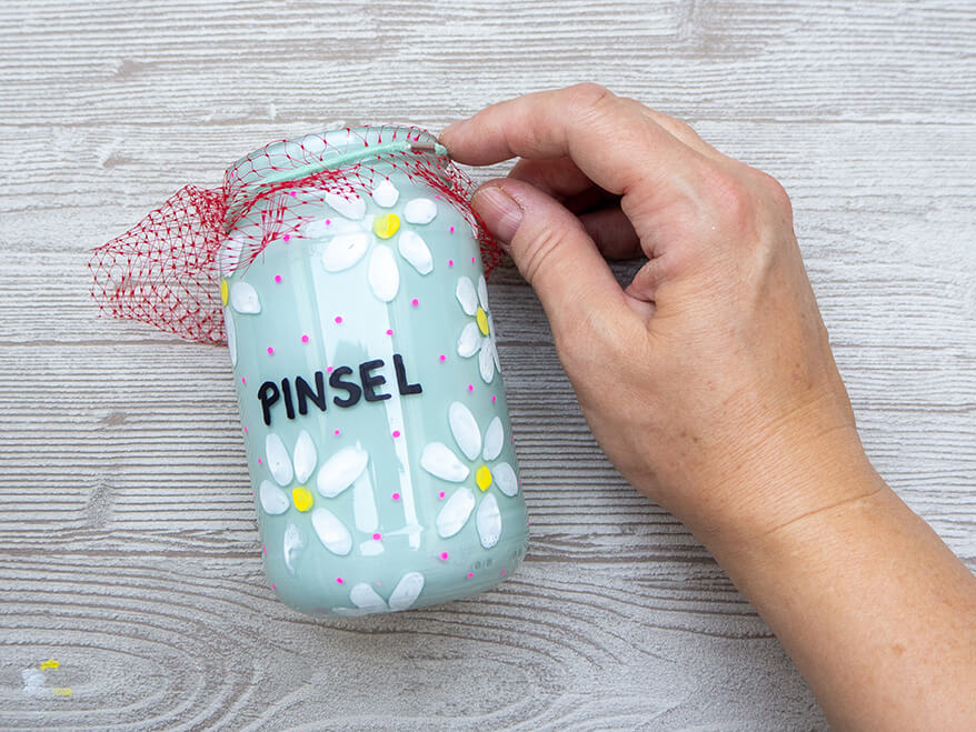 Jemand zieht auf einem hellgrauen Holzuntergrund über ein leeres Marmeladenglas, das von innen mit hellblauer Acrylfarbe ausgesprüht wurde und mit Acrylmarkern angemalt wurde, ein rotes Netz.