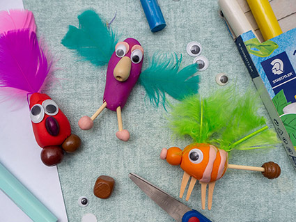 Auf einem türkisfarbenen Untergrund liegen drei selbstgebastelte Fantasiefiguren aus Knete. Sie wurden mit Federn, Perlen, Wackelaugen und Zahnstochern verziert. Eines ist rot, eines violett und das dritte orange. Drumherum sind verschiedene Bastelmaterialien von Staedtler verteilt. Foto: Johanna Rundel