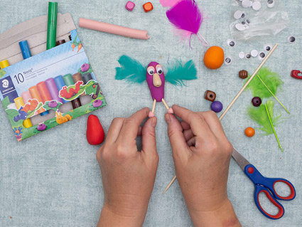 Auf einem türkisfarbenen Untergrund bastelt jemand eine Fantasiefiguren aus lila Knete von Staedtler und verziert sie mit Federn, Perlen, Wackelaugen und Zahnstochern. Drumherum sind verschiedene Bastelmaterialien verteilt. Foto: Johanna Rundel