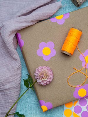 Auf einem türkisfarbenen Untergrund liegt ein in Kraftpapier eingepacktes Geschenk. Darauf sind mit pinken und neonorangen Markierungspunkten von Avery Zweckform kleine Blumen geklebt. Auf dem Geschenk liegen eine Rolle neonoranges Bäckergarn und eine rosafarbene Dahlienblüte. Unter dem Geschenk liegen weitere Aufkleberbögen mit Markierungspunkten. Daneben ist ein rosa Tuch drapiert. Foto: Johanna Rundel