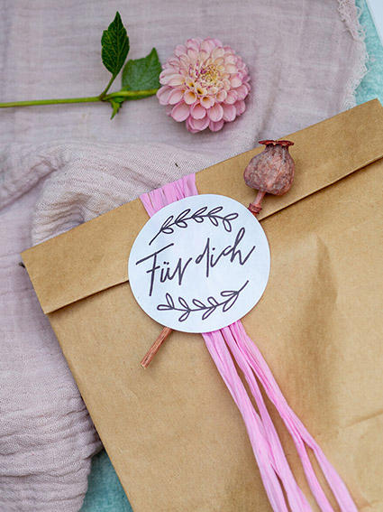 Auf einer rosa Tischdecke liegt eine braune Papiertüte um die ein rosa Band gewickelt wurde. Darauf wurde ein runder Aufkleber von Avery Zweckform mit der Aufschrift "Für dich" geklebt. Unter dem Aufkleber klemmt eine pinke Trockenblume. In der linken, oberen Ecke des Bildes liegt eine rosafarbene Dahlienblüte. Foto: Johanna Rundel