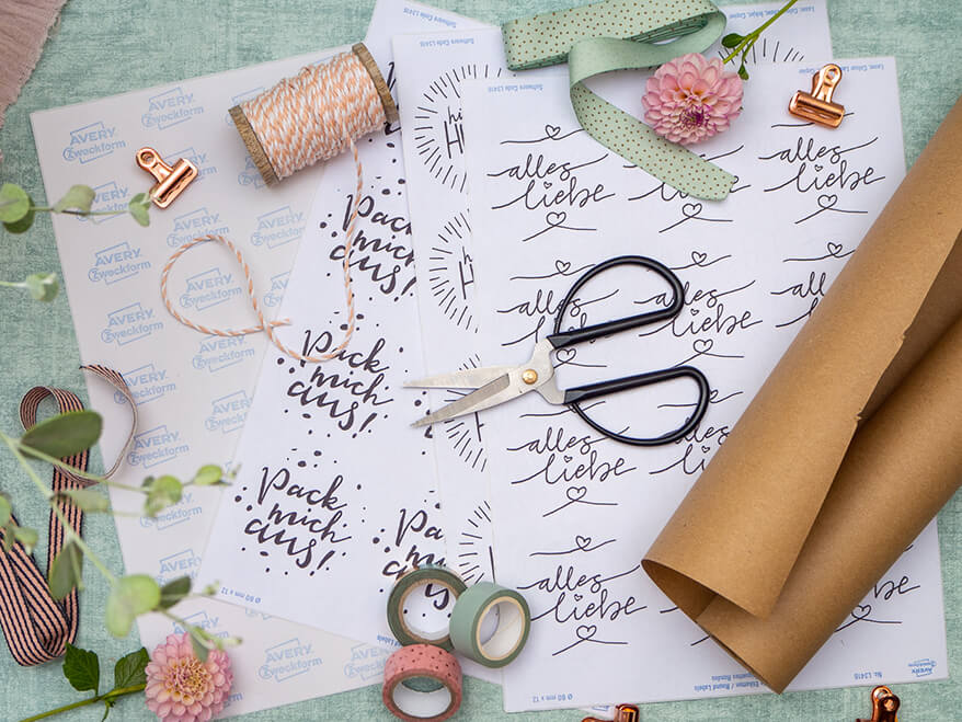 Auf einem türkisfarbenen Untergrund liegen mehrere runde Blanko-Etikettbögen von Avery Zweckform verteilt, die mit unterschiedlichen Geschenkaufkleber-Designs bedruckt wurden. Auf den Bögen liegt eine schwarze Schere, eine Rolle Bäckergarn, diverse Geschenkbänder, eine Rolle Packpapier, kupferfarbene Klemmen und verschiedene Washitapes. Einzelne rosa Dahlienblüten lockern das Bild auf. Foto: Johanna Rundel