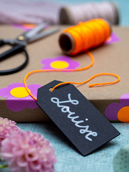 Auf einem türkisfarbenen Untergrund liegt ein in Kraftpapier eingepacktes Geschenk. Darauf sind mit pinken und neonorangen Markierungspunkten von Avery Zweckform kleine Blumen geklebt. Auf dem Geschenk liegt eine Rolle neonoranges Bäckergarn und eine schwarze Schere. An das Geschenk gelehnt ist ein schwarzes Tafeletikett, auf dem mit weißer Schrift „Louise“ steht. Im Vordergrund liegen zwei rosa Dahlienblüten. Foto: Johanna Rundel
