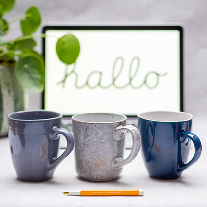 Auf einem hellgrauen Untergrund stehen drei Tassen in verschiedenen Blautönen. Im Vordergrund liegt ein orangener Drehgriffel von Leuchtturm1917. Im Hintergrund steht ein Laptop, auf dessen Bildschirm "hello" steht. Links daneben steht eine Zimmerpflanze. Foto: Johanna Rundel 