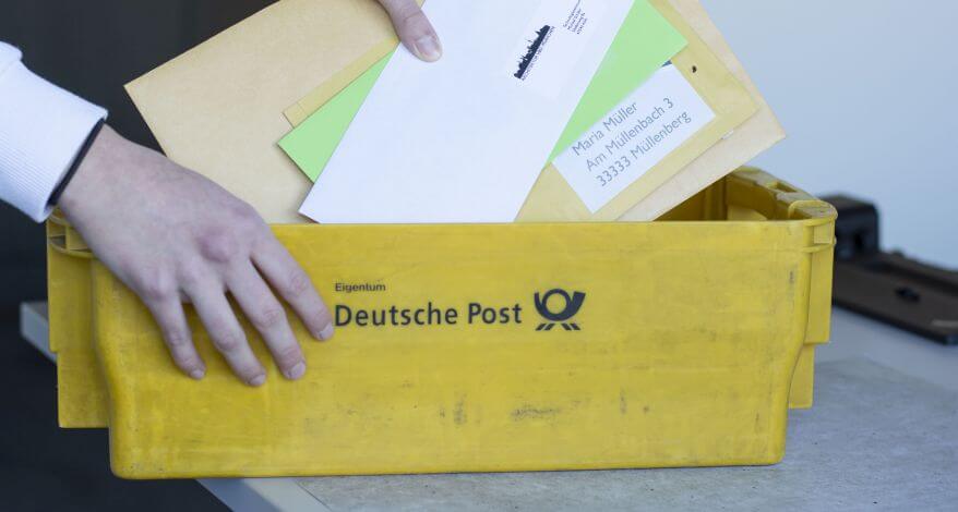 Eine große gelbe Postbox mit dem Schriftzug und Logo der Deutschen Post steht auf einem Tisch. Eine Hand hält die Box fest, die andere steckt Briefe und Versandtaschen, die mit Adressetiketten von Avery Zweckform beklebt sind, in die Postbox.