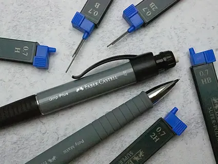 Auf einem hellgrauen Untergrund liegen zwei dunkelgraue Druckbleistifte von Faber-Castell. Drumherum sind die grauen Behälter aus Plastik mit den verschiedenen Ersatzminen verteilt. Sie haben blaue Deckel. Foto: Johanna Rundel
