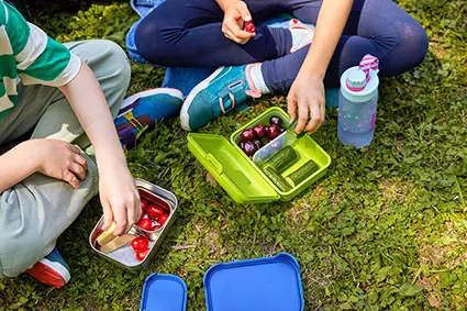 alt="Zwei Grundschüler sitzen im Schneidersitz auf dem Gras und essen Obst und Gemüse aus ihren Brotboxen von ergobag."