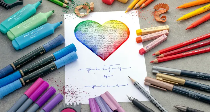 Viele bunte Stifte der Marke Faber-Castell liegen am Rand eines Blatt Papiers, auf das ein Herz gemalt wurde.