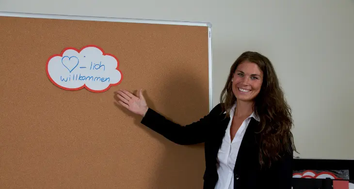 Das Bild zeigt eine lächelnde Frau, die auf eine Moderationstafel aus Kork zeigt. Auf der Tafel ist eine beschriftete Wolkenkarte mit der Aufschrift „Herzlich willkommen“ angebracht. 