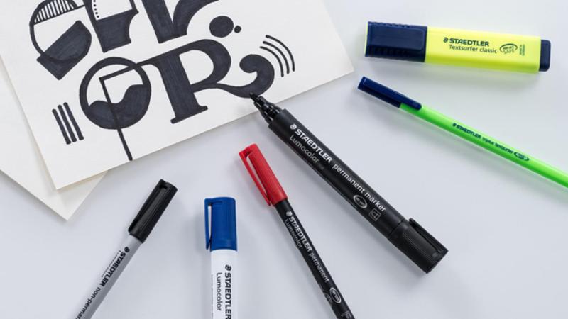 Diverse farbige Marker von Staedtler und ein gelettertes Papier liegen auf einem Tisch