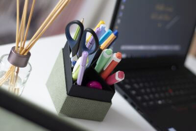 Ein gefüllter Stifteköcher der S.O.H.O. Kollektion von Rössler steht auf einem Schreibtisch. Im Hintergrund steht ein aufgeklappter Laptop. Neben dem Stifteköcher steht ein Lufterfrischer