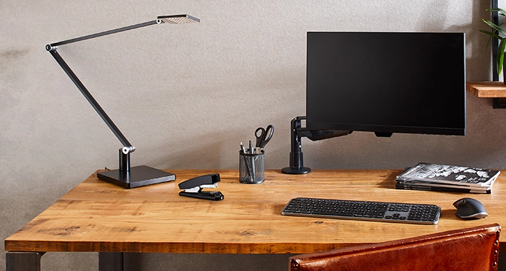 Auf einem aufgeräumten Holzschreibtisch befinden sich eine Schreibtischlampe, ein Monitor, eine Tastatur, eine Maus, ein Novus-Heftgerät sowie ein Stiftehalter mit Scheren und Stiften. Im Hintergrund sind eine Wand, ein Regal und eine Pflanze zu sehen. 