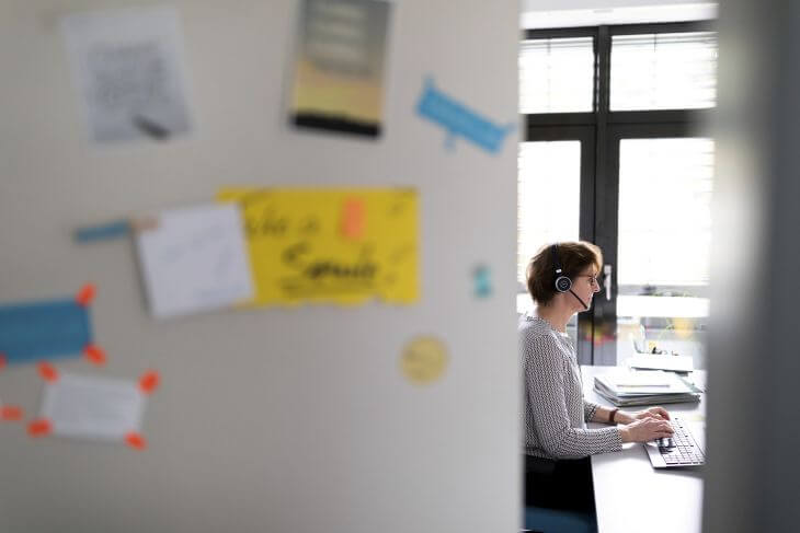 Eine Tür zu einem Büro ist verschwommen im Bild. Durch einen Spalt sieht man eine Frau mit Headset an einer Tastatur sitzen und arbeibten.